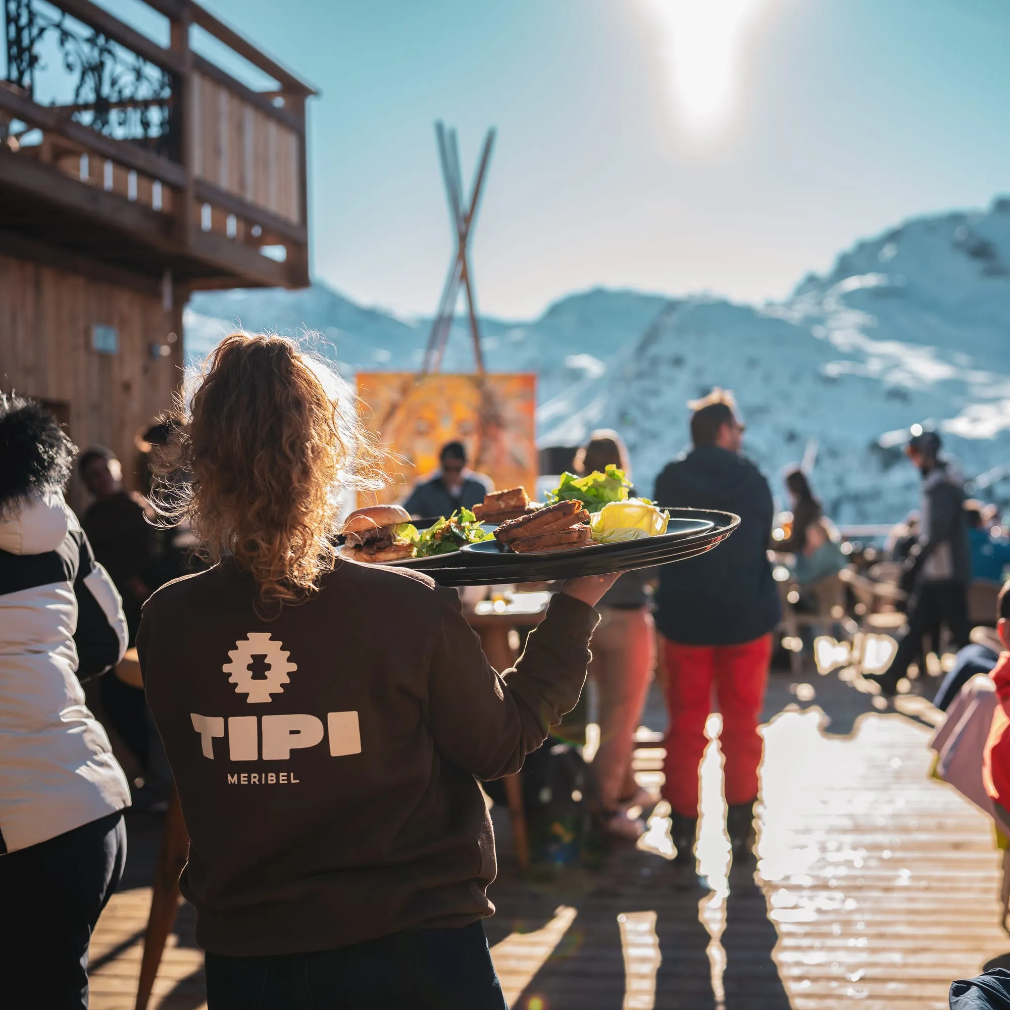 Une serveuse en sweat Tipi porte les plats sur la terrasse, au soleil.